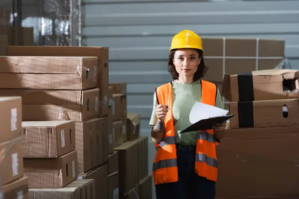 Mujer trabajadora de almacén en casco duro sujetando portapapeles y mirando a la cámara, gestión logística - foto de stock