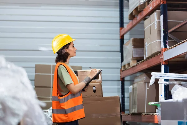 Side view, female warehouse worker in hard hat and safety vest holding clipboard checking inventory — Stock Photo