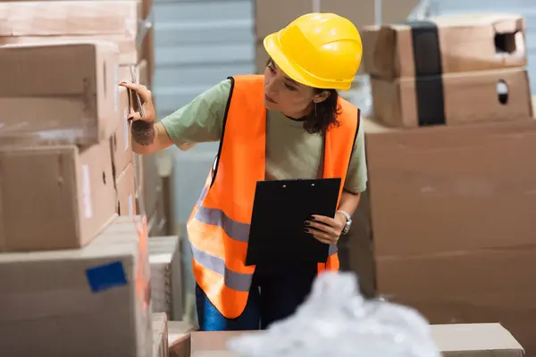 Female warehouse worker in hard hat and safety vest holding clipboard while checking cargo — Stock Photo