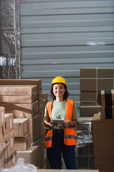 Веселая женщина-работница склада в бронежилете и каске с цифровым планшетом рядом с грузом — стоковое фото