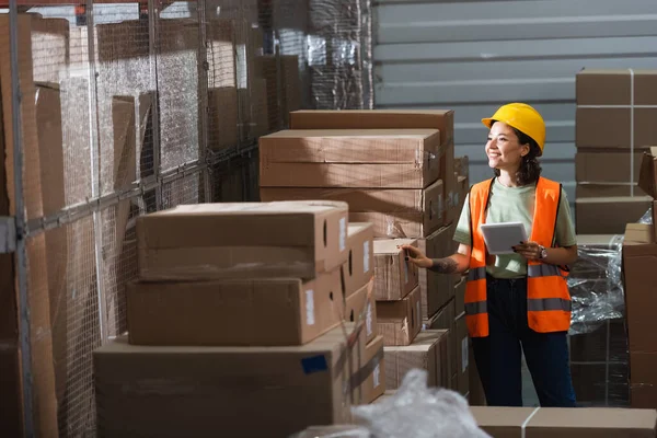 Happy female warehouse worker in safety vest and hard hat holding digital tablet near cargo — Stock Photo