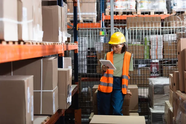 Female warehouse worker in safety vest and hard hat holding digital tablet near cargo boxes — Stock Photo
