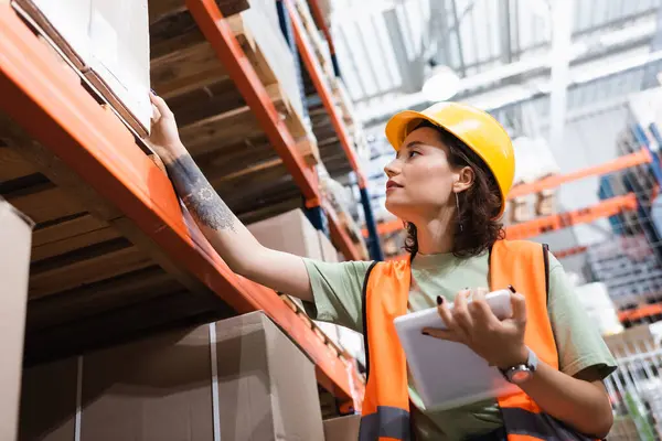 Tattooed female warehouse worker in safety vest and hard hat holding digital tablet near cargo boxes — Stock Photo