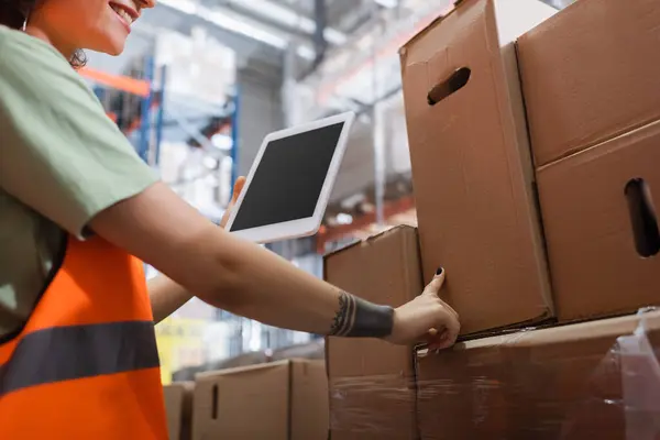 Cropped female warehouse worker in safety vest smiling and holding digital tablet and checking cargo — Stock Photo