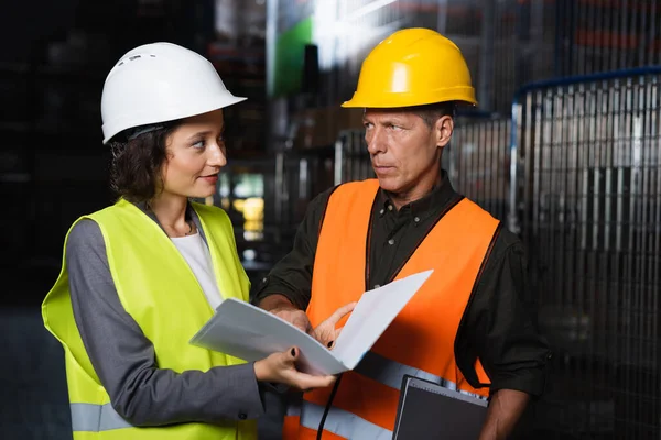 Two warehouse workers discussing logistics while standing together in hard hats and safety vests — Stock Photo