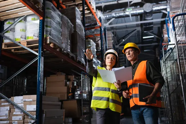 Warehouse workers discussing logistics, woman with folder showing direction to middle aged colleague — Stock Photo