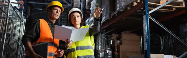 Warehouse workers banner, woman with folder showing direction to middle aged colleague — Stock Photo