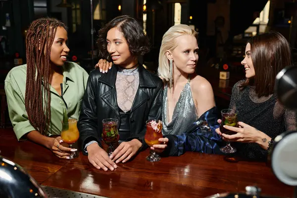 Novias multiétnicas alegres y elegantes con copas de cóctel en el bar, vibrante vida nocturna - foto de stock