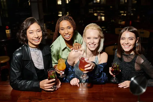 Charming and happy multiethnic girlfriends with cocktail glasses looking at camera in bar — Stock Photo