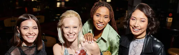 Elegant and happy multiethnic girlfriends with cocktail glasses smiling at camera in bar, banner — Stock Photo