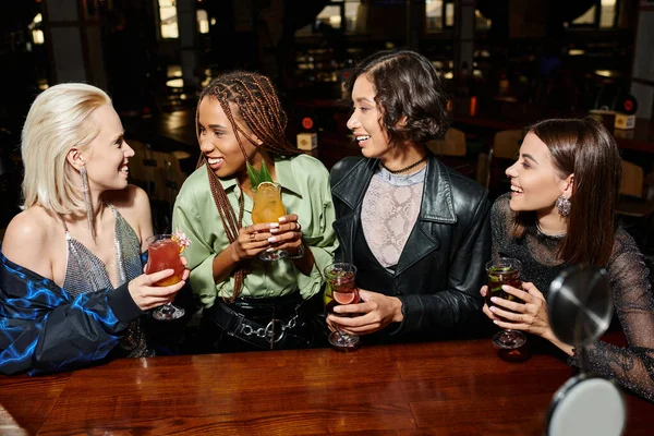 Smiling and stylish multiracial girlfriends with cocktail glasses talking in bar, vibrant atmosphere — Stock Photo