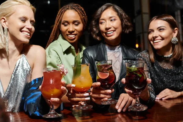 Amigas multiculturales felices y encantadoras celebrando deliciosos cócteles en el bar, fiesta glamorosa - foto de stock