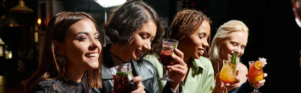Щасливі і стильні мультикультурні подружки тримають смачні коктейлі в нічному барі, банер — стокове фото