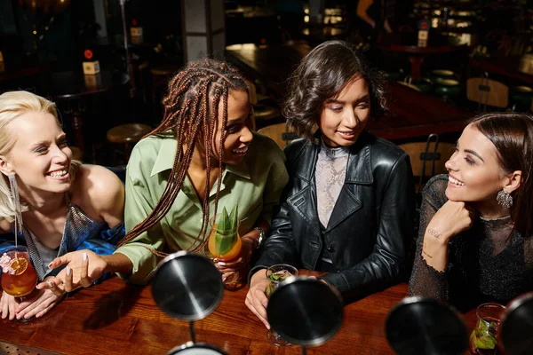 Joyeuses amies multiethniques parlant et souriant près de verres à cocktail, fête de poule dans le bar — Photo de stock