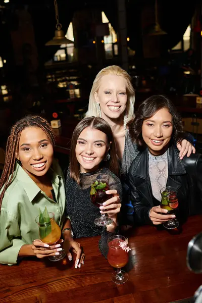Модные и счастливые многонациональные подруги, держащие вкусные коктейли и смотрящие в камеру в баре — стоковое фото