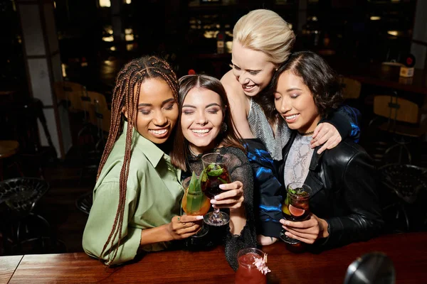 Joyful multicultural girlfriends clinking cocktail glasses and smiling with closed eyes in bar — Stock Photo