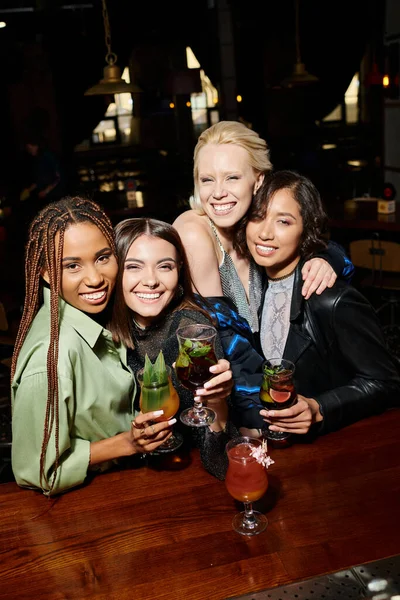 Sorridenti fidanzate multietniche con bicchieri da cocktail che abbracciano e guardano la fotocamera nel bar — Foto stock