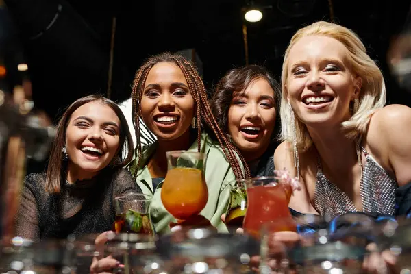 Веселые многорасовые подружки, смотрящие в камеру рядом с вкусными коктейлями в баре, досуге и веселье — стоковое фото