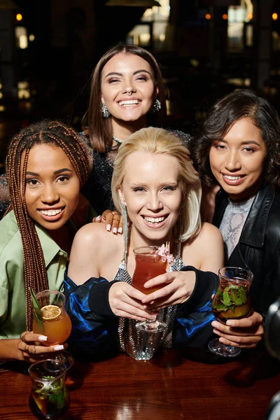 Веселые и гламурные мультикультурные женщины с коктейлями, смотрящие в камеру в баре, праздничная вечеринка — стоковое фото