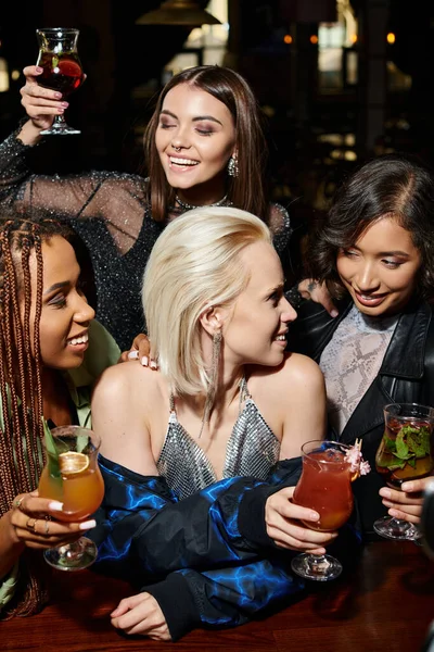 Улыбающиеся многорасовые элегантные женщины с вкусными коктейлями, веселящиеся в баре, развлекающиеся в ночное время — стоковое фото