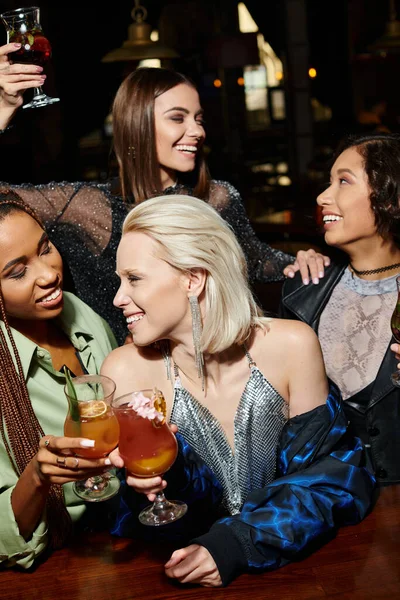 Happy alluring women clinking cocktail glasses near multiethnic girlfriends during party in bar — Stock Photo