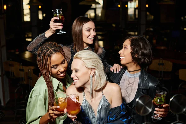 Cheerful elegant women clinking cocktail glasses near multiethnic girlfriends during party in bar — Stock Photo
