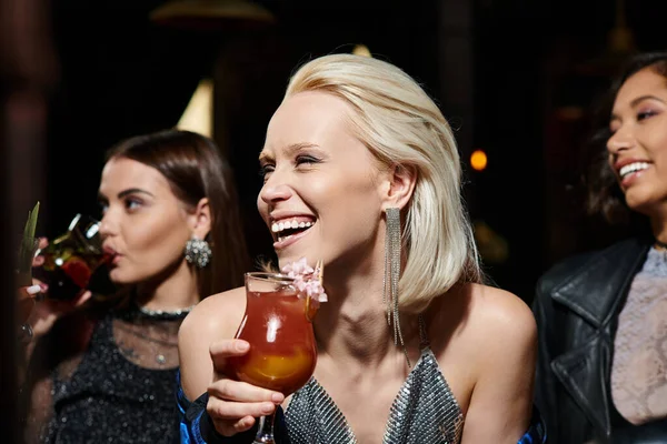 Cheerful blonde woman holding cocktail glass near multiethnic girlfriends during hen party in bar — Stock Photo
