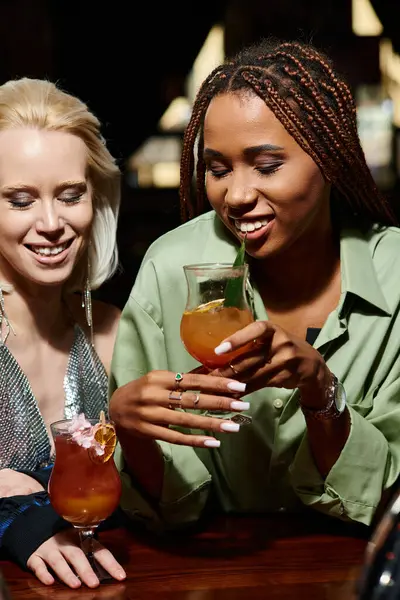 Novias multirraciales jóvenes y glamorosas sosteniendo vasos con cócteles en el bar, tiempo de fiesta - foto de stock