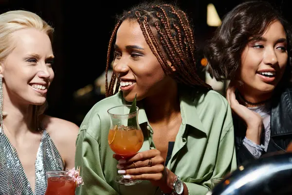 Gaie femme afro-américaine avec verre à cocktail souriant près de copines multiethniques dans le bar — Photo de stock