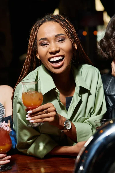 Mulher americana africana animado e na moda com copo de coquetel rindo perto de namoradas no bar — Fotografia de Stock