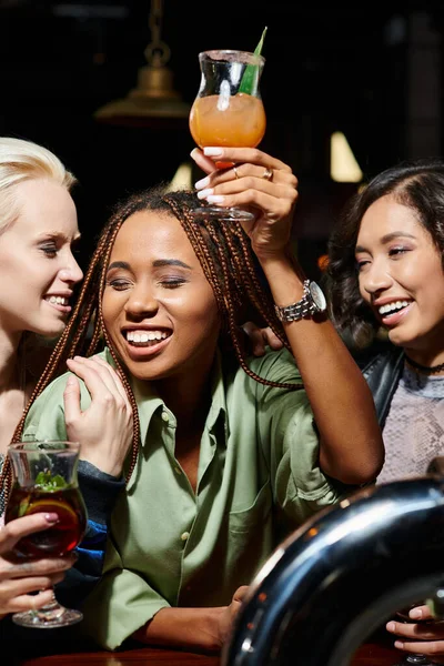 Joyeuse femme afro-américaine toastant avec verre à cocktail près de copines multiethniques élégantes — Photo de stock