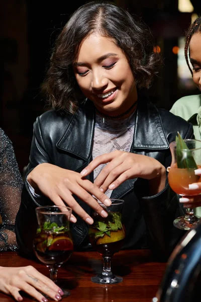 Jeune et élégant asiatique femme souriant près de verre à cocktail et copines multiculturelles dans le bar — Photo de stock