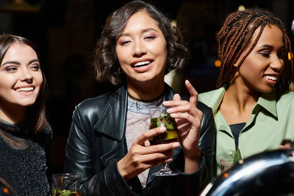 Молода і модна азіатка з коктейль-окулярами посміхається біля багатоетнічних подруг в нічному барі — стокове фото