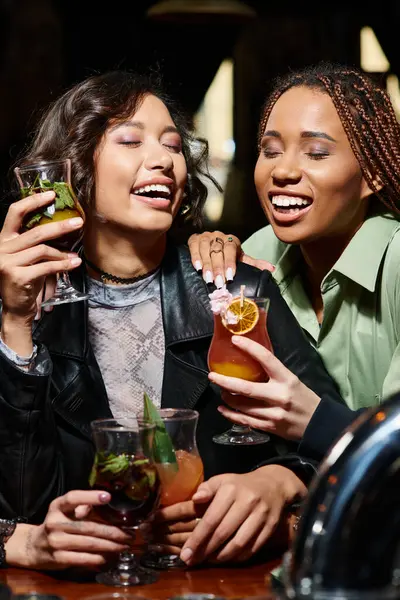 Mujeres afroamericanas y asiáticas emocionadas sosteniendo cócteles y riendo con los ojos cerrados en el bar - foto de stock