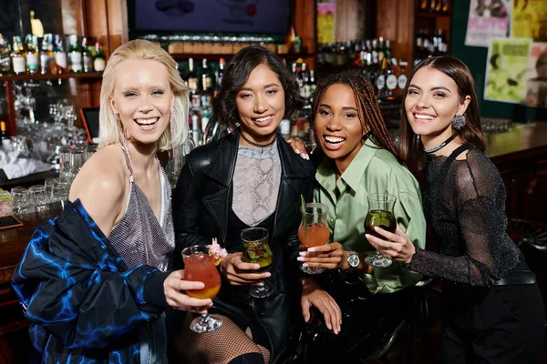 Веселые и элегантные многонациональные девушки в коктейльных очках, смотрящие в камеру в баре — стоковое фото