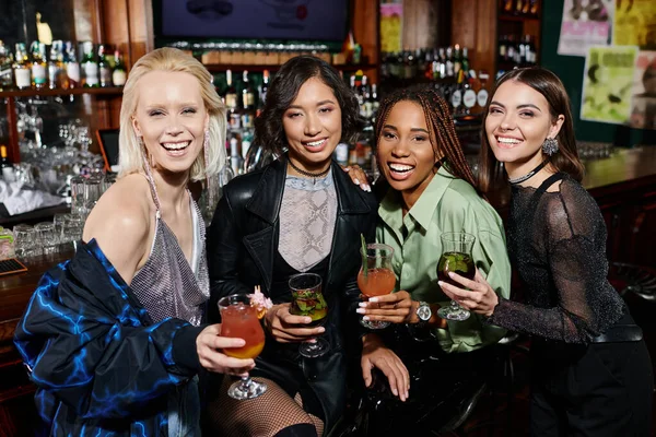 Cheerful and elegant multiethnic girlfriends with cocktail glasses looking at camera in bar — Stock Photo