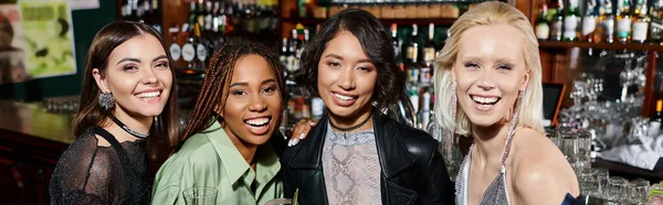 Joyful and stylish multiethnic girlfriends with cocktail glasses looking at camera in bar, banner — Stock Photo