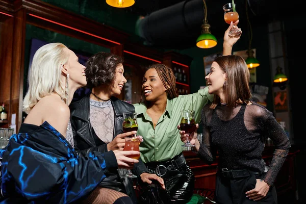 Excité femme afro-américaine toasting avec cocktail près de copines multiethniques élégantes dans le bar — Photo de stock