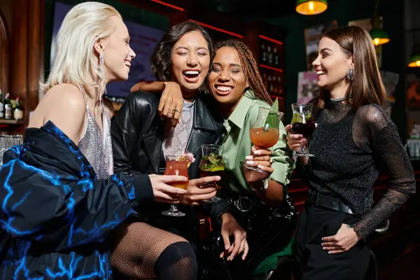 Alegres novias multiétnicas con copas de cóctel abrazando y riendo en el bar, emoción - foto de stock