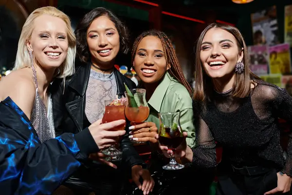 Стильные и счастливые мультиэтнические женщины звонят в коктейльные очки и улыбаются в баре перед камерой — стоковое фото