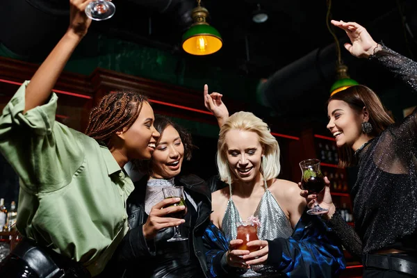 Excited and trendy multicultural girlfriends with cocktails glasses dancing in bar, glamorous party — Stock Photo