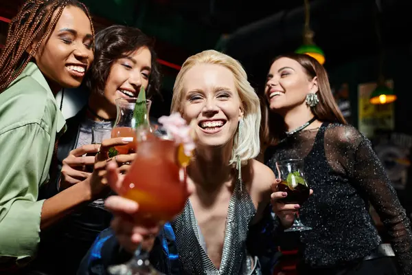 Heureuse femme blonde avec un verre de cocktail souriant à la caméra près de copines multiethniques dans le bar — Photo de stock