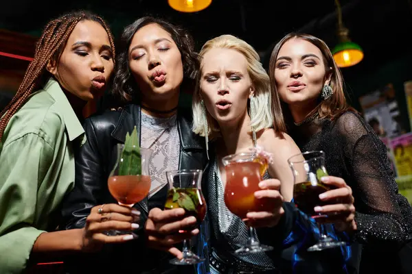 Гламурные многонациональные девушки в коктейльных очках, дующиеся губами, веселясь в баре — стоковое фото