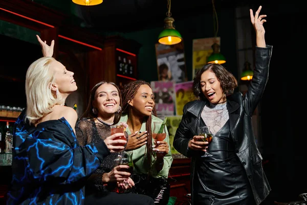 Очаровательные и счастливые многонациональные девушки танцуют с коктейльными бокалами в баре, гламурная вечеринка — стоковое фото