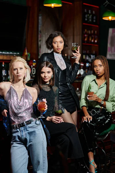 Confident and classy multiethnic girlfriends with cocktail glasses looking at camera in bar, party — Stock Photo