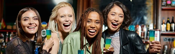 Stylish multiracial girlfriends with shot glasses laughing and looking at camera in bar, banner — Stock Photo
