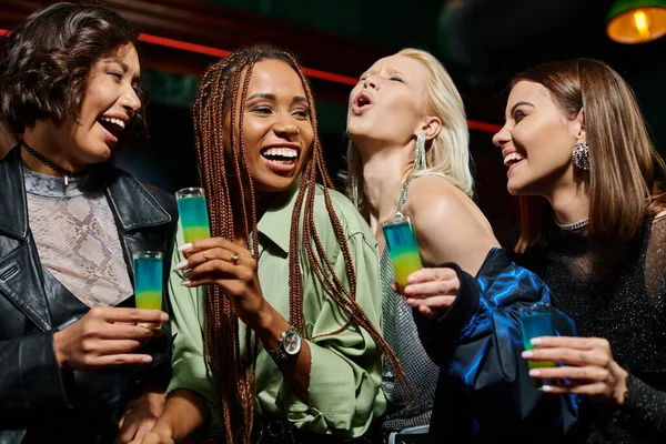 Overjoyed and cheerful multicultural girlfriends holding shot glasses in bar, festive party — Stock Photo