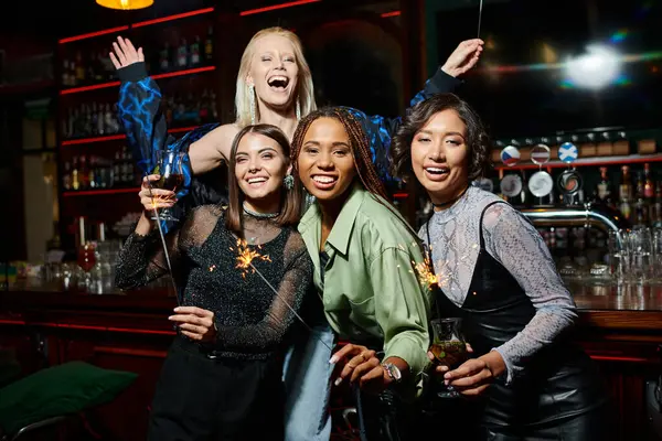 Joyful multiethnic girlfriends with cocktails and festive sparklers having fun in bar, festive party — Stock Photo