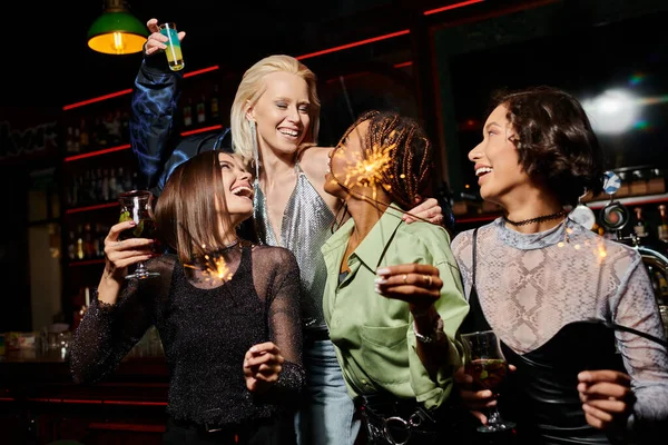 Novias multiétnicas felices y elegantes con cócteles y chispas festivas durante la fiesta en el bar - foto de stock