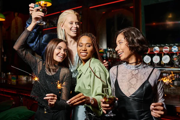 Joyful multiethnic girlfriends toasting with cocktails and holding shiny sparklers in bar, party — Stock Photo